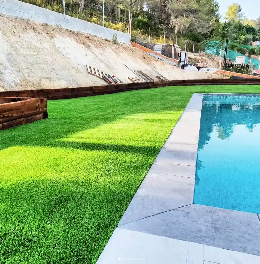 Colocación de césped artificial drenante alrededor de una piscina con acabados de madera en el Penedès.